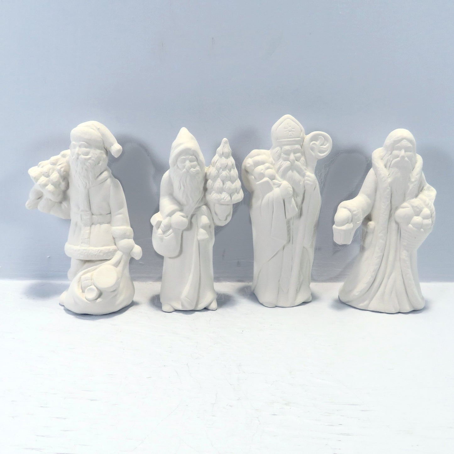 Set of 4 handmade ready to paint ceramic santa figurines on a pale blue surface. They are facing forward carrying various items