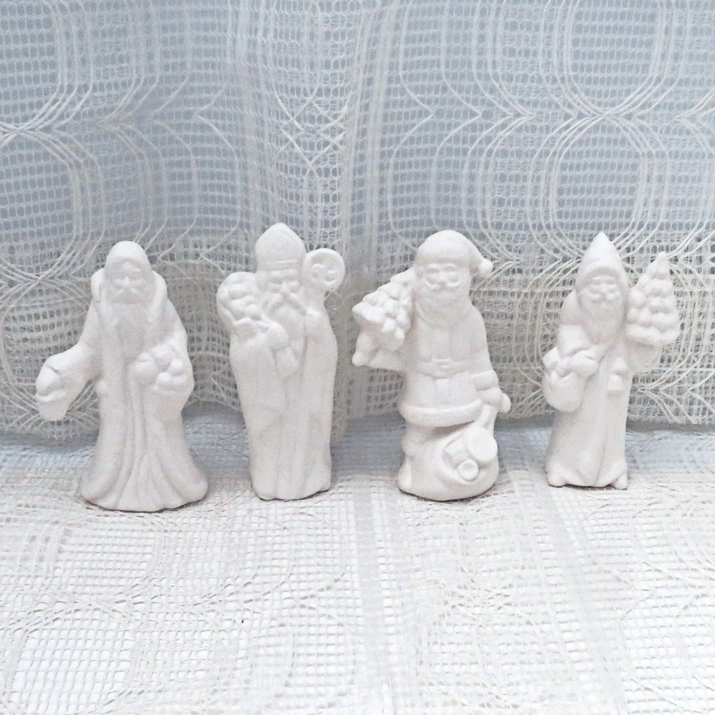 4 ready to paint ceramic Santas on a lacy ecru table cloth with a curtain behind them.