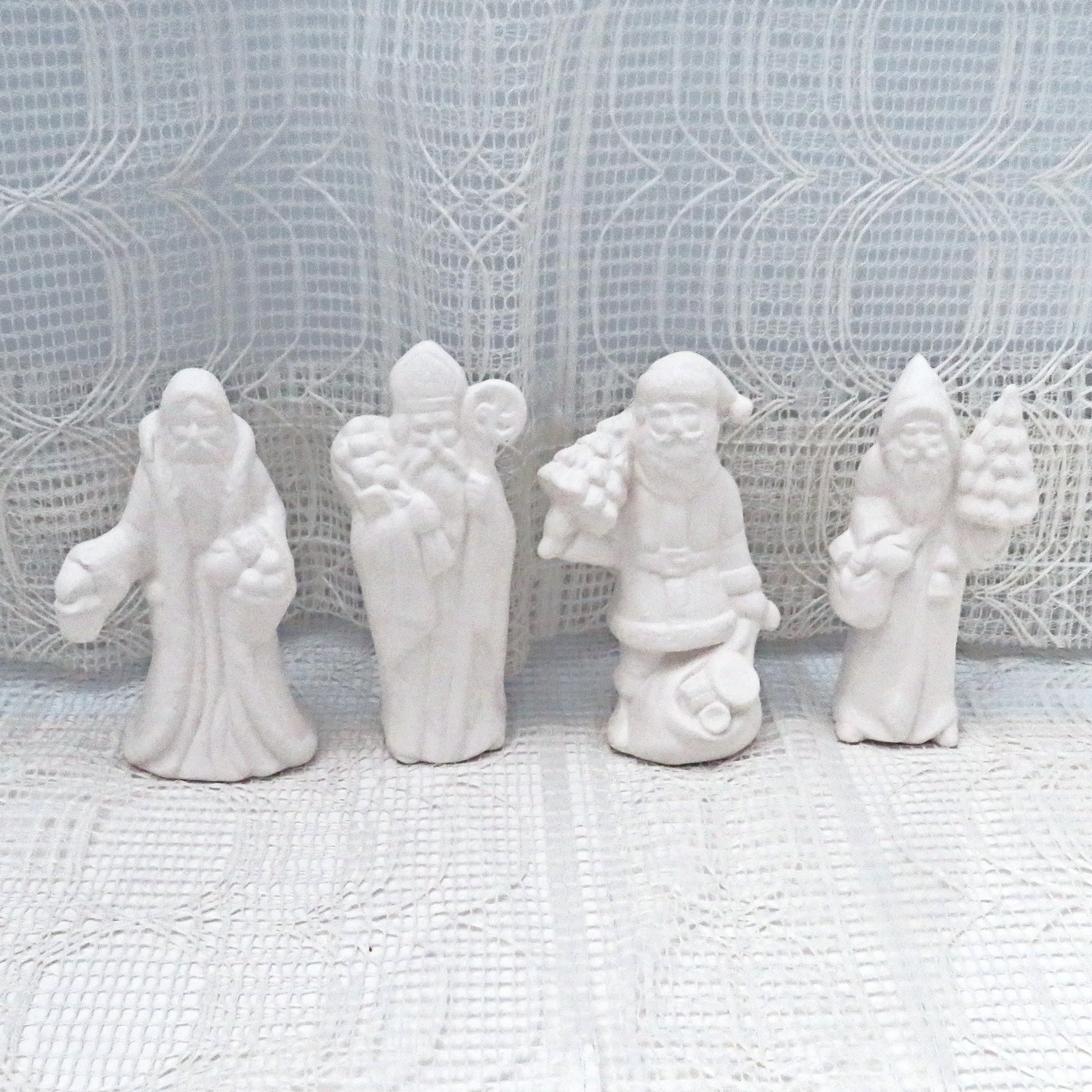 4 ready to paint ceramic Santas on a lacy ecru table cloth with a curtain behind them.