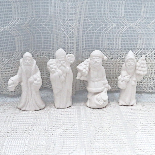 4 ready to paint ceramic Santas on a lacy ecru table cloth with a curtain behind them.