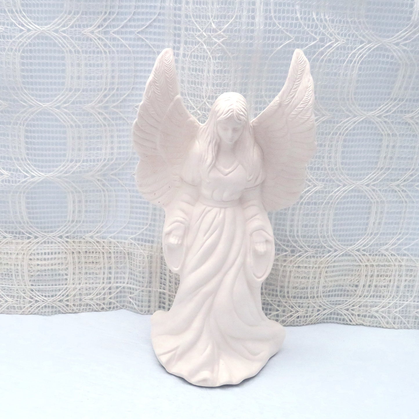 Unpainted ceramic angel standing on a pale blue table with an ecru curtain in the background. Her wings are up, her hands are forward and at her side. Her head is slightly bowed.