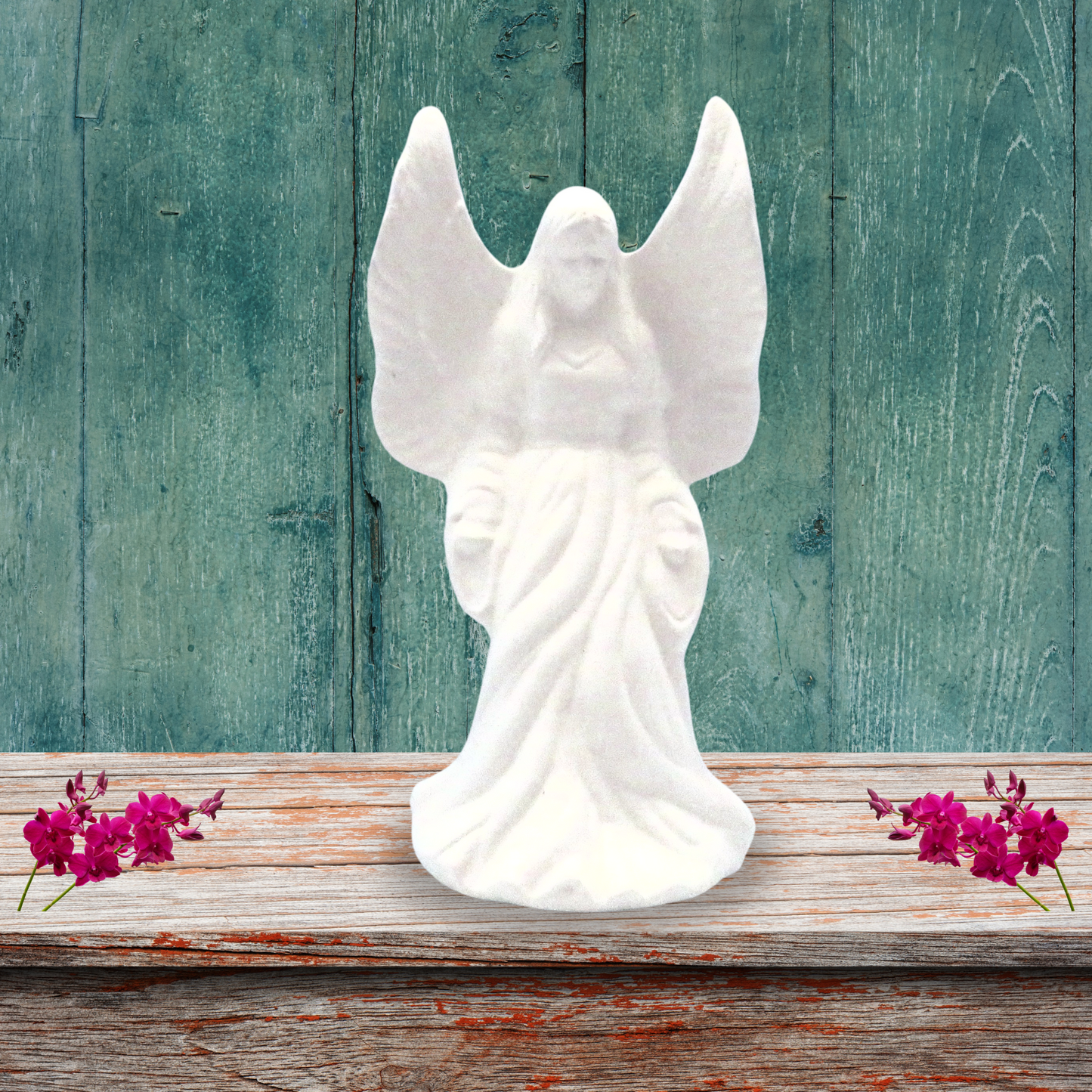 Handmade unpainted ceramic standing angel figurine standing on a rustic brown shelf with a dusty green background. There are small pink flowers on the shelf on either side of her.