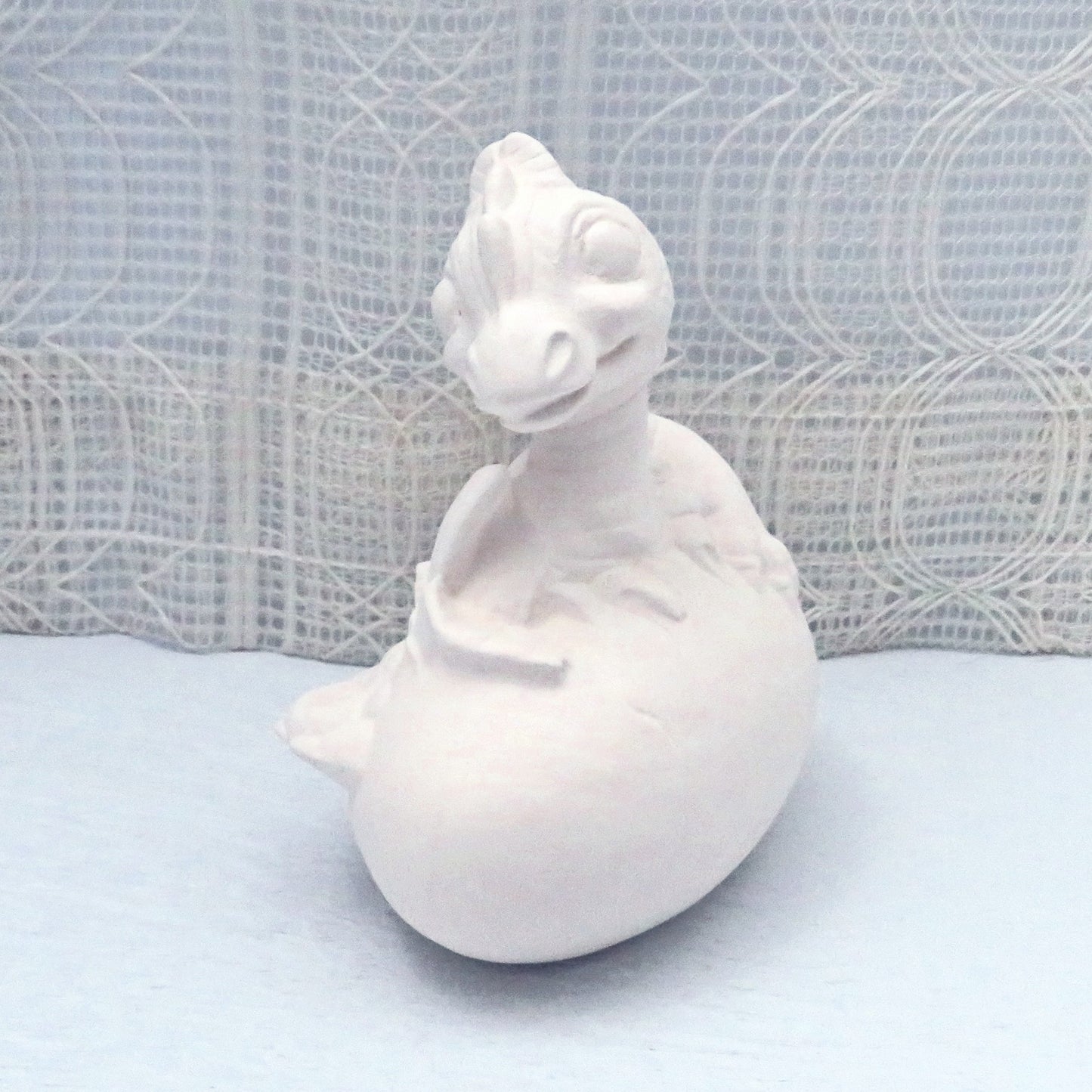 Side View of unpainted ceramic dragon coming out of her egg. Her face is looking at the camera.