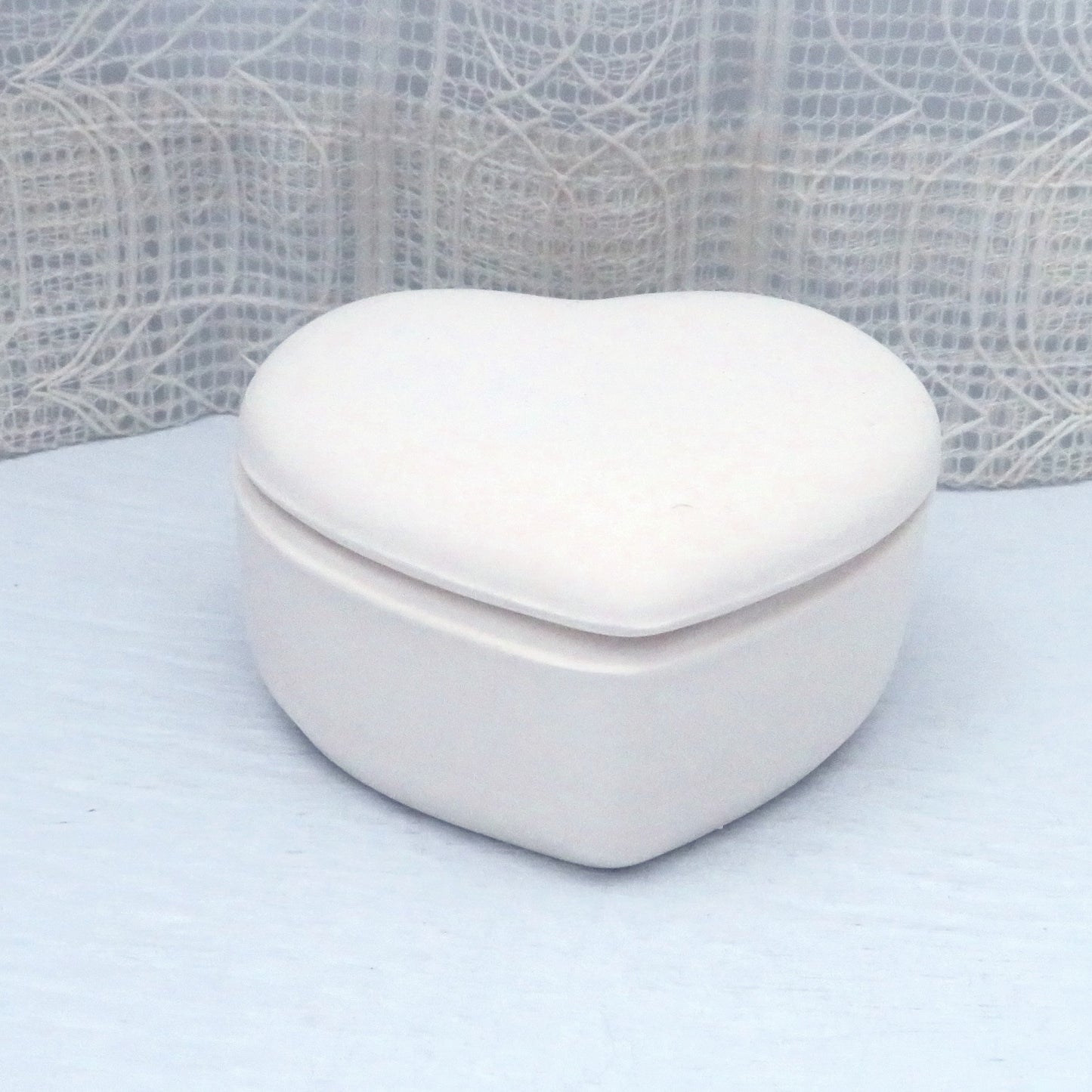 Paintable ceramic heart lidded dish on a pale blue table with an ecru lacy curtain behind it.