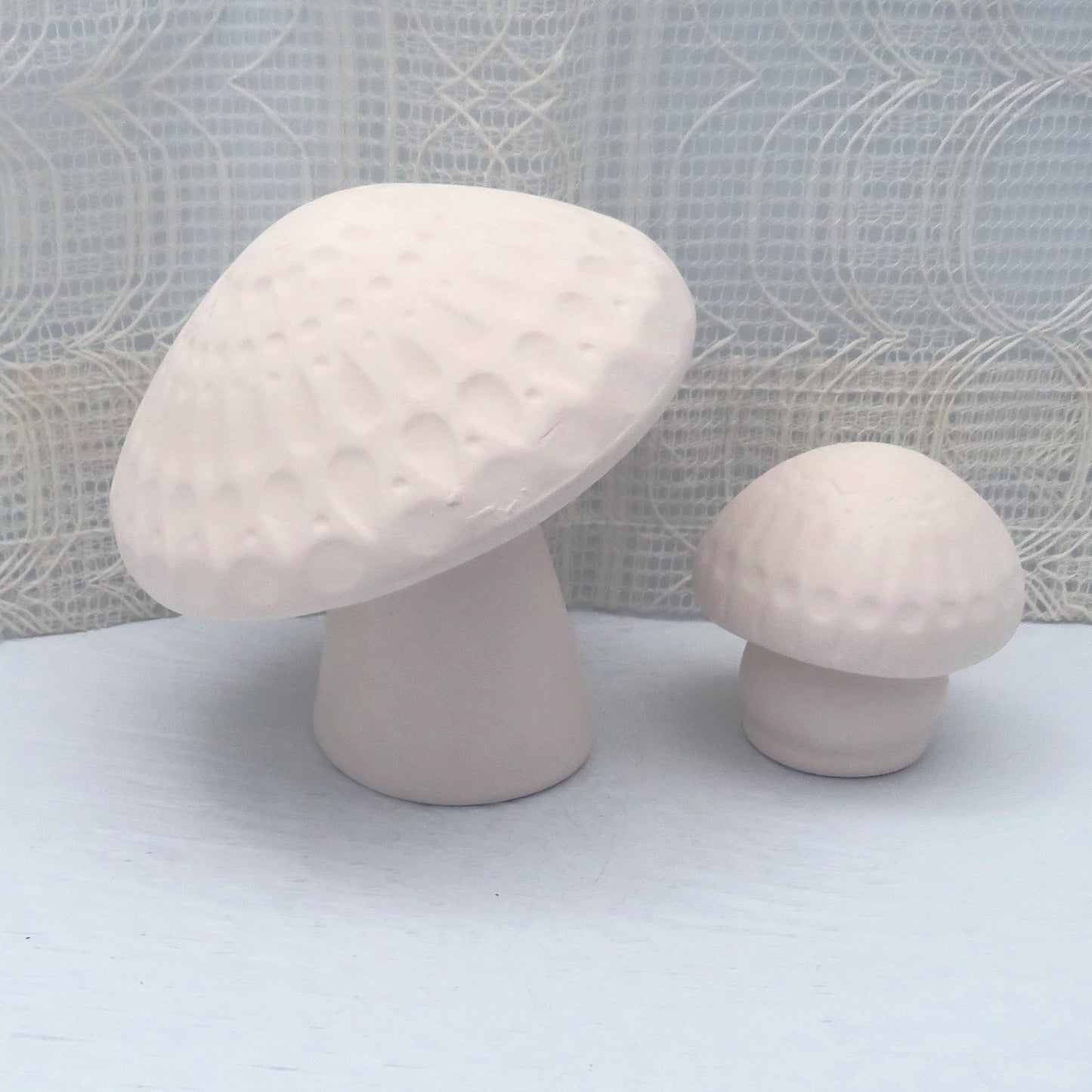 Paintable ceramic mushroom statues on a blue table with an ecru lacy curtain behind it. You can see and feel the texture in the tops of the mushrooms. One mushroom is larger, one smaller.