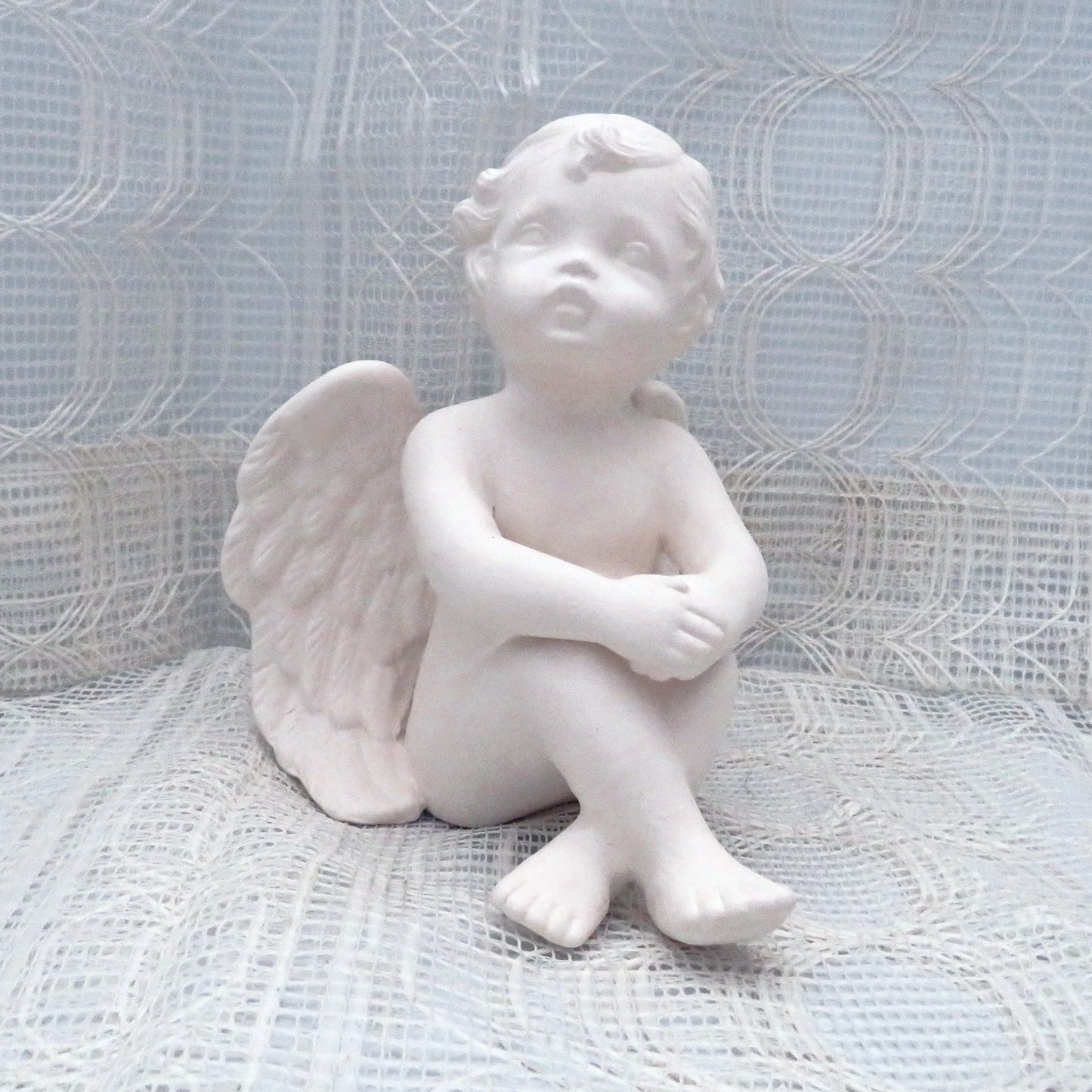 6 inch paintable ceramic cherub statue sitting on a lacy ecru table cloth with a curtain behind it. His knees are up and his hands are resting on his knees.
