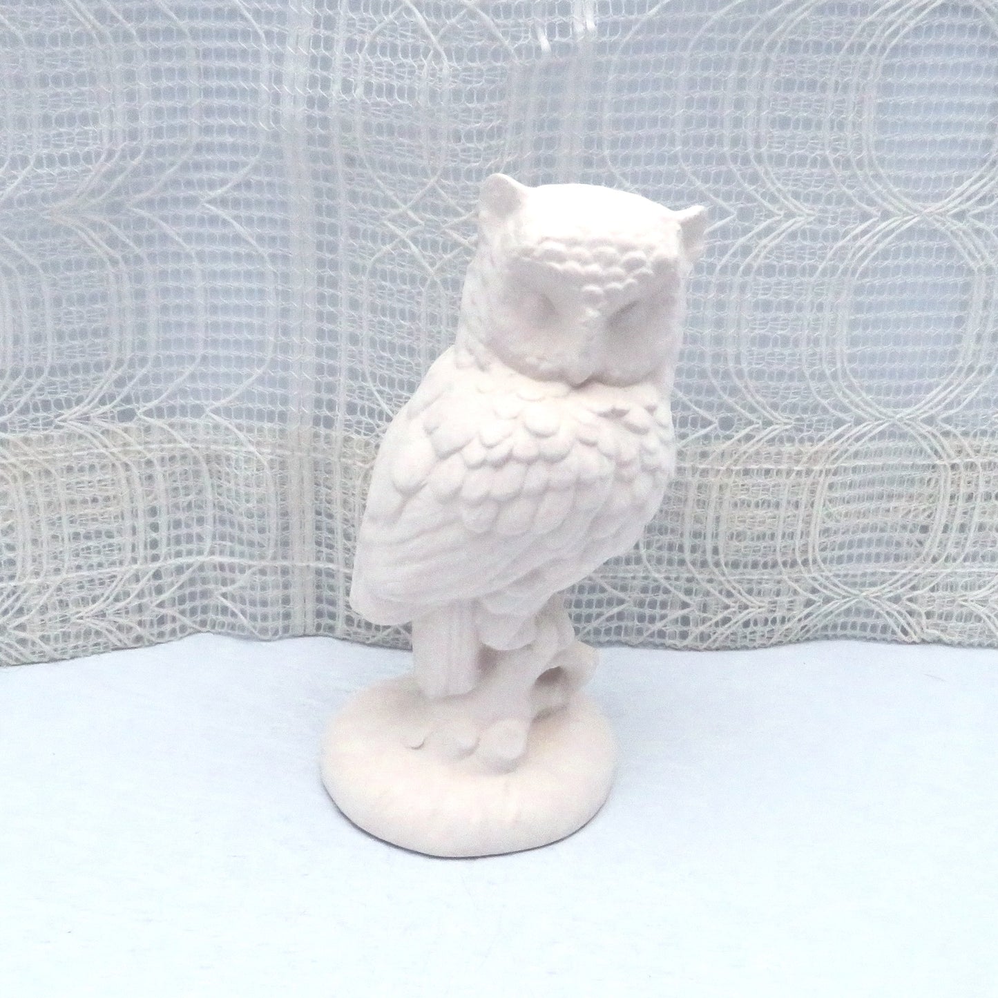 Ready to paint ceramic owl figurine sitting on a pale blue table with an ecru lacy curtain behind. The owl is and looks like it si standing on a tree stump.