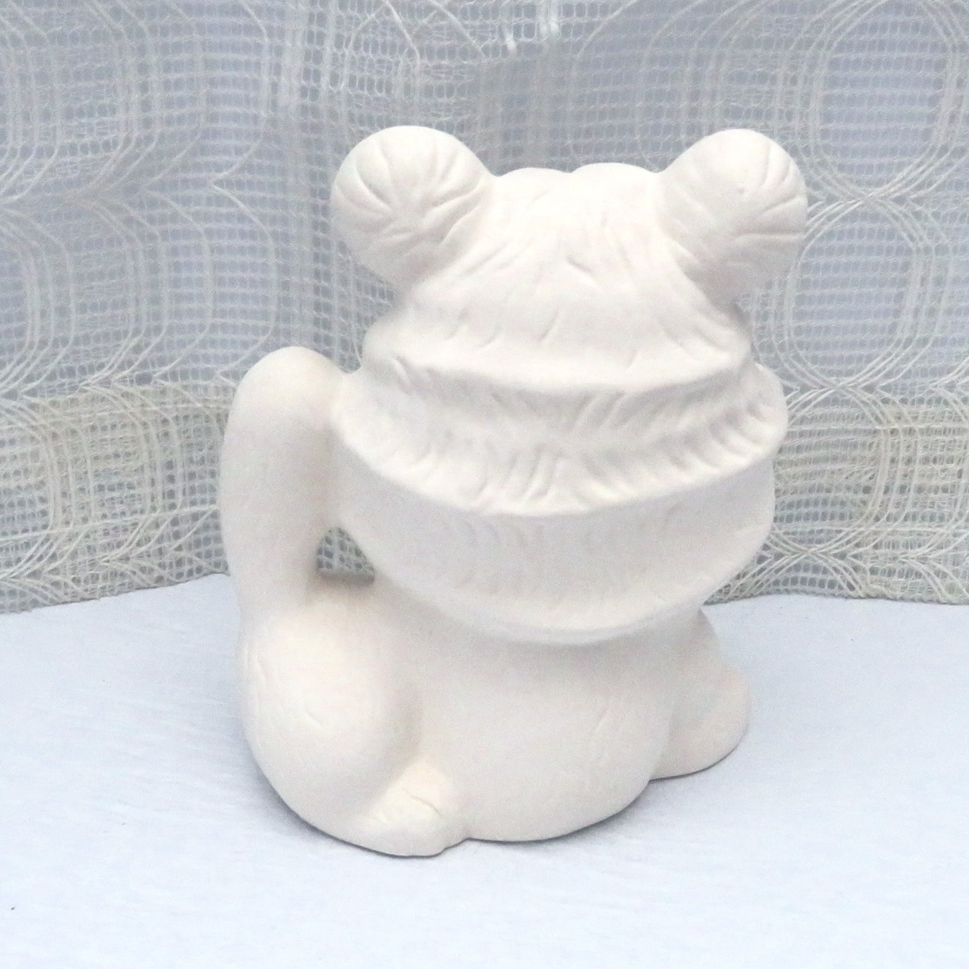 Unpainted ceramic tiger statue facing away from the camera.  It is on a pale blue table with a lacy ecru curtain behind it.