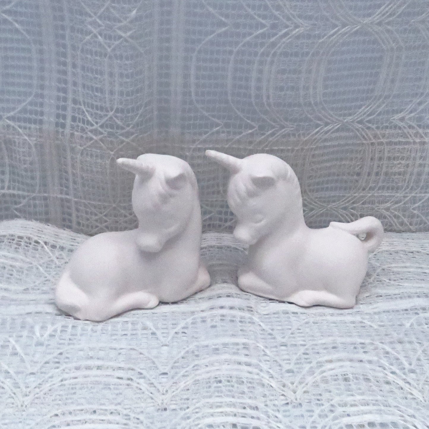 2 paintable unicorn statues head to head on an ecru lacy curtain.