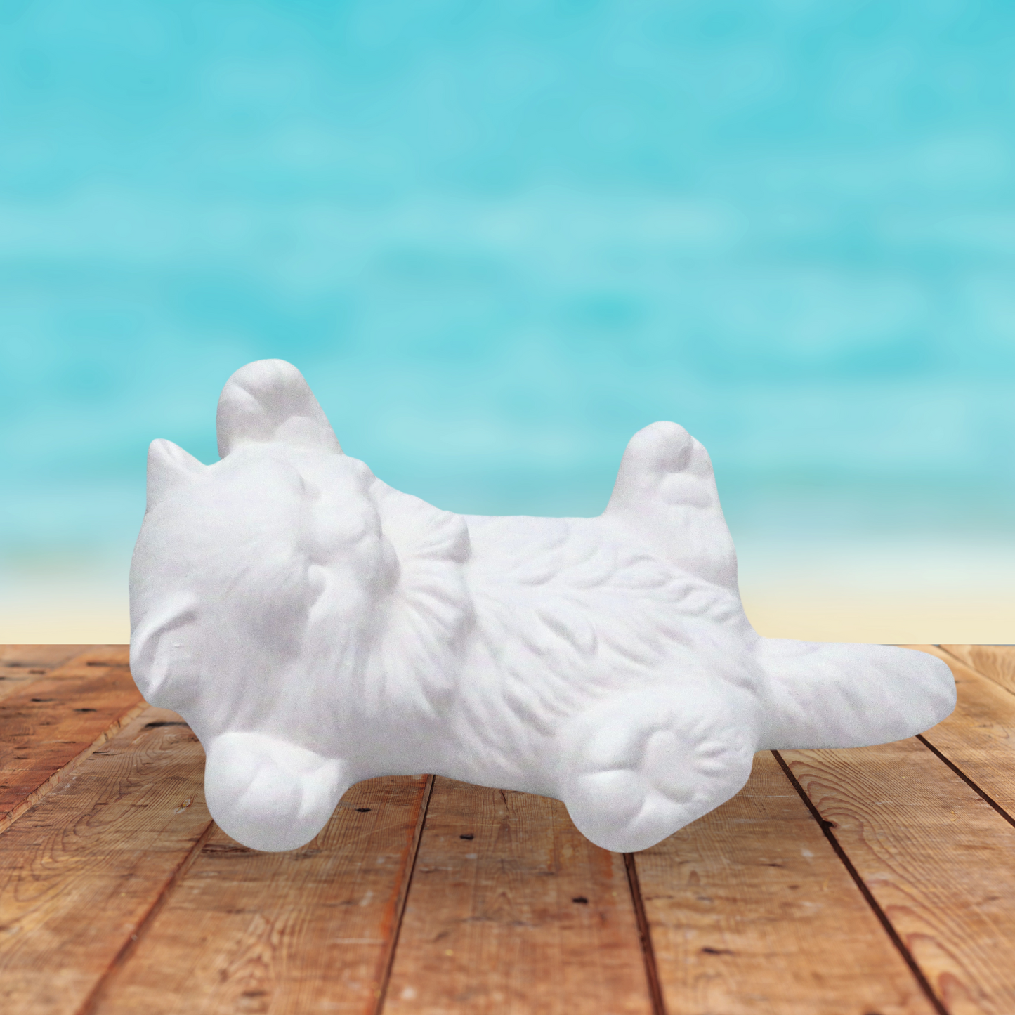 Handmade Ready to Paint ceramic kitten on its back with its legs out and its tail straight down. It's lying on a rustic table by the ocean.