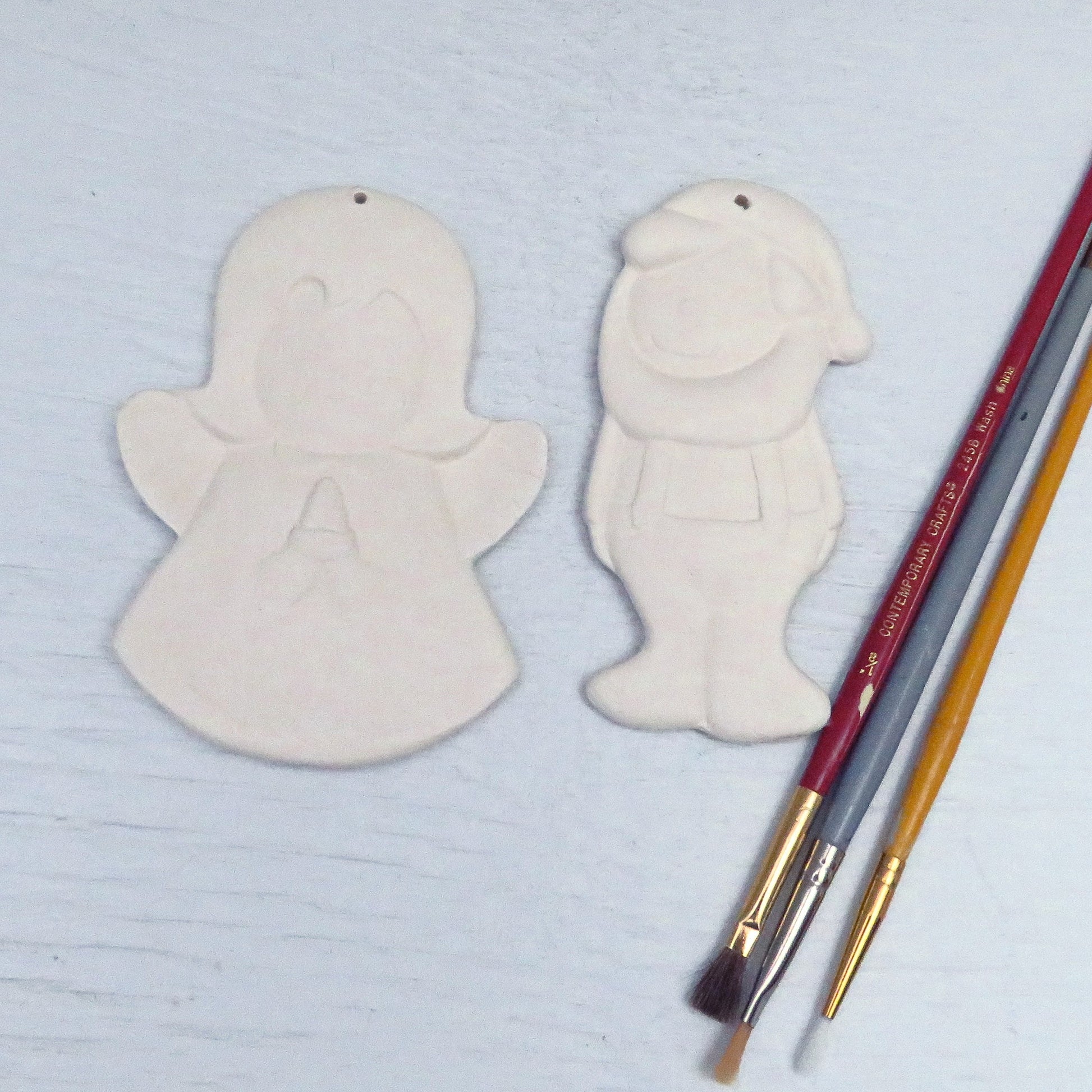 ready to paint ceramic angel and elf ornaments on a pale blue background with 3 paint brushes on the right.