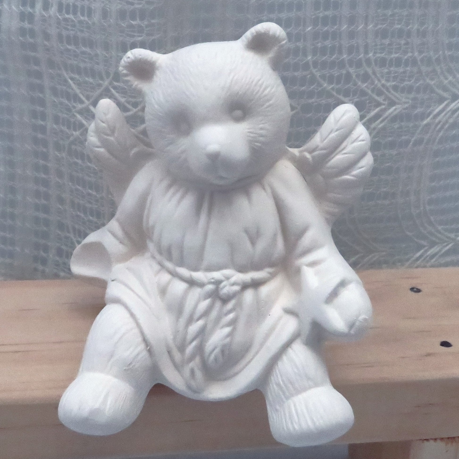 Close up view of unpainted ceramic bear angel sitting on a wooden bench.