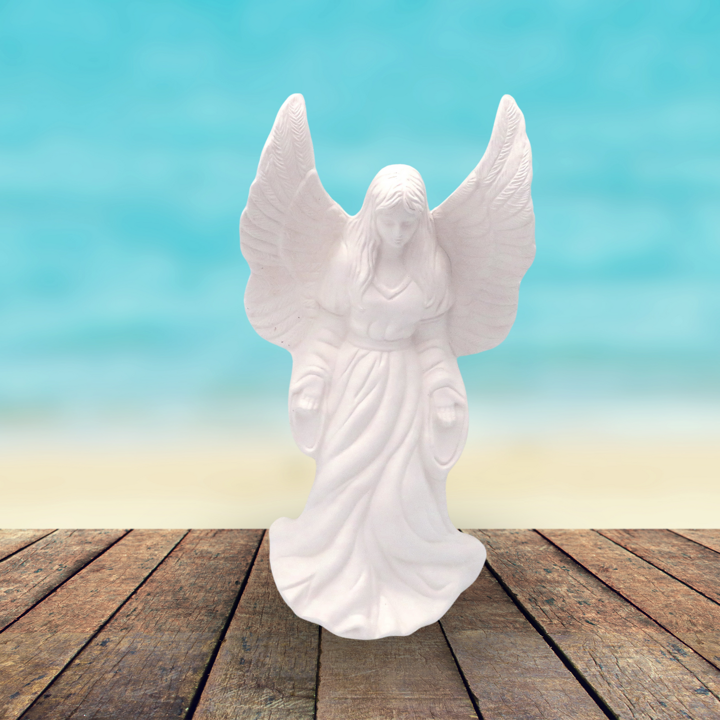 Handmade Ready to Paint ceramic angel statue standing on rustic brown wooden table by a smooth ocean background. Her wings are outstretched, her head is slightly bowed. Her arms are by her side and hands facing forward.