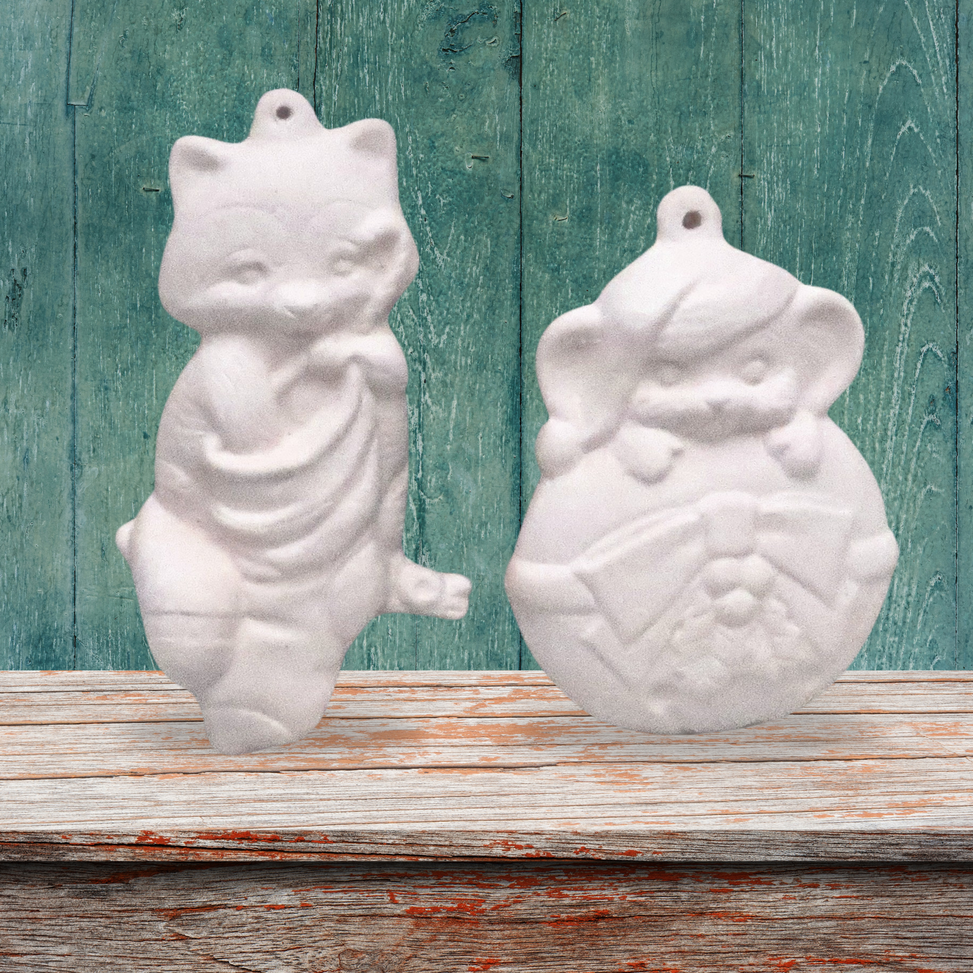 set of 2 ready to paint ceramic christmas tree ornaments on a wood shelf with a green background.  One ornament is a cat with his hand in the stocking.  The other is a mouse climbing over an ornament.