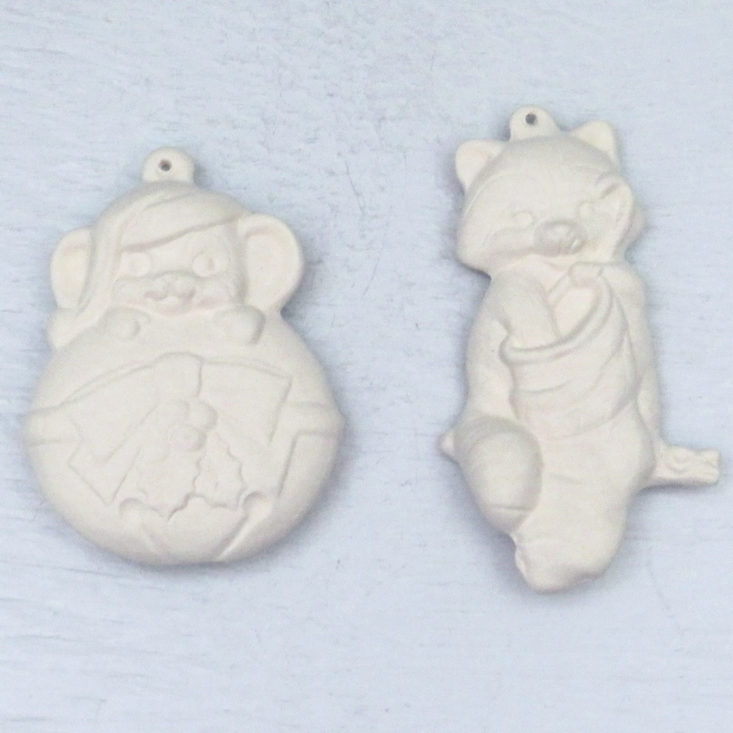 ready to paint cat ornament with its hand inside a stocking. Unpainted ceramic mouse ornament hanging on to a round Christmas tree ornament. They are on a pale blue surface.