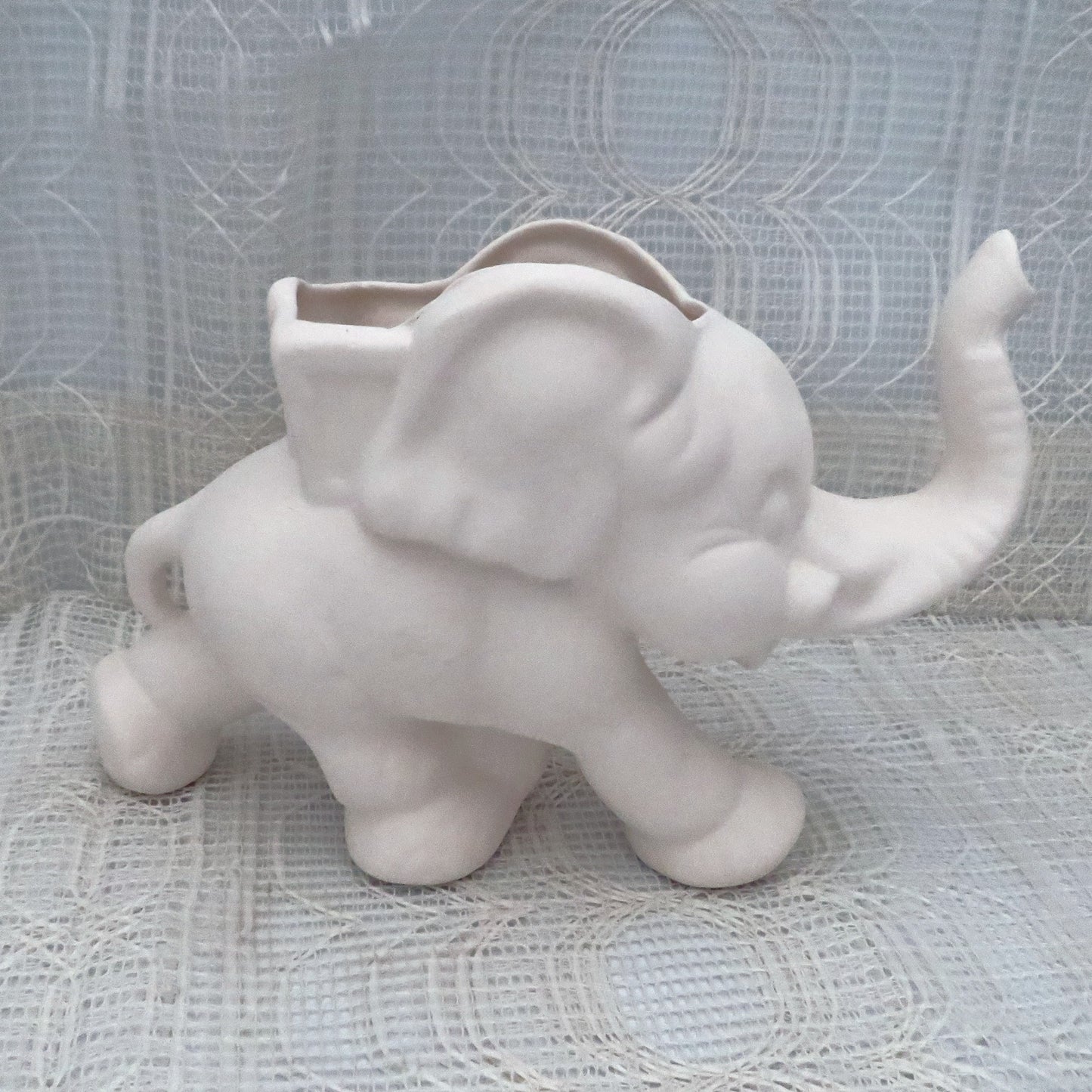 Unpainted ceramic elephant statue on an ecru table cloth with a curtain behind it.  It's trunk is in the air, and it is in a running position.