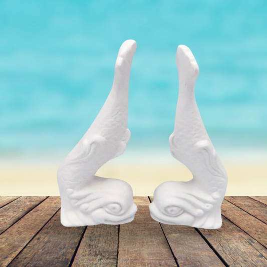 1 set of 2 ready to paint ceramic fish statues sitting on a rustic wooden table by the ocean.