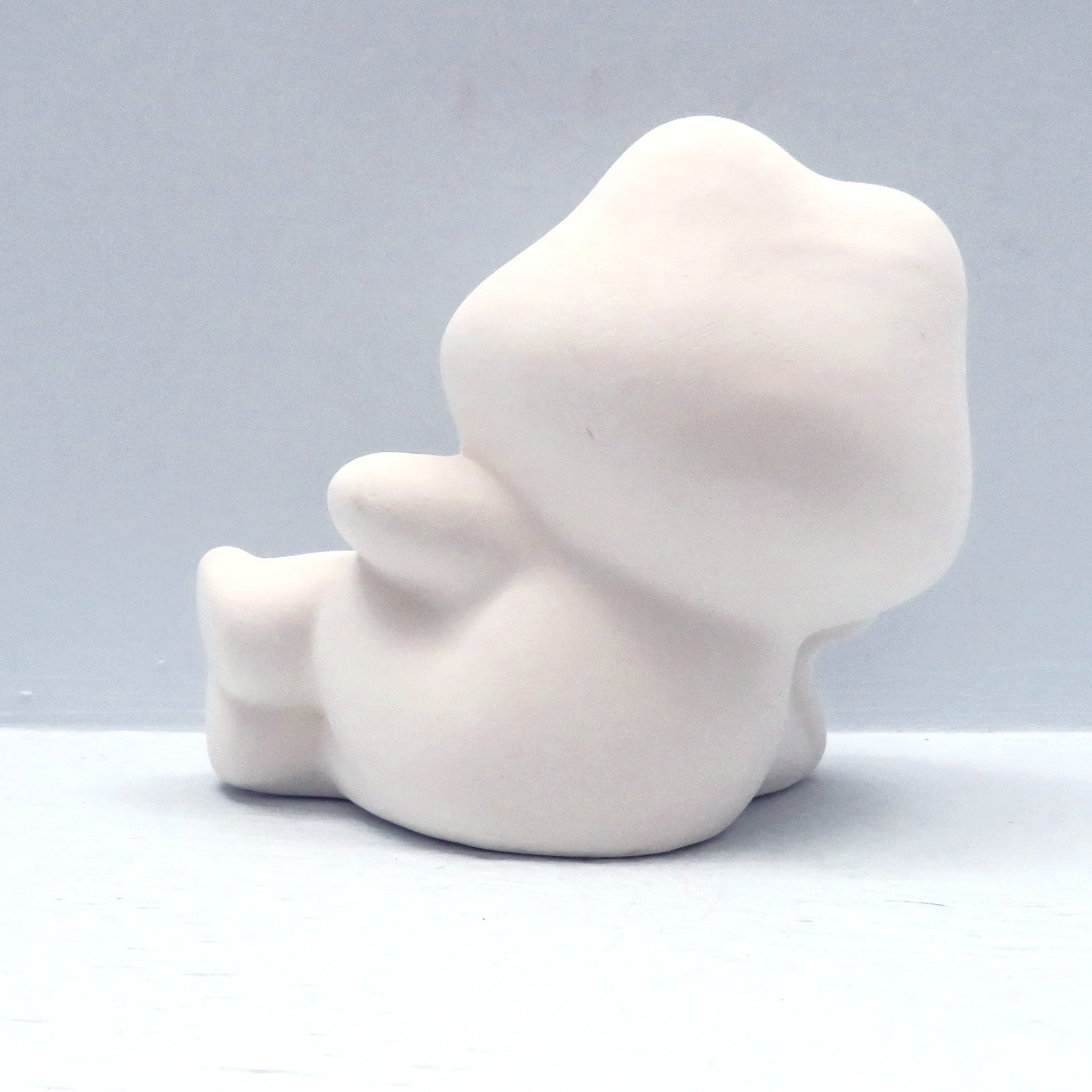 Handmade paint it yourself ceramic frog on his side.  He is facing away from the camera and lying on a pale blue background.