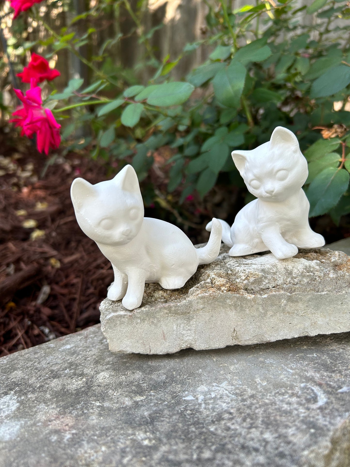 2 handmade sitting ready to paint cats sitting on white stones in front of a rose bush. the cats are sitting wth their tails near each other and their faces forward