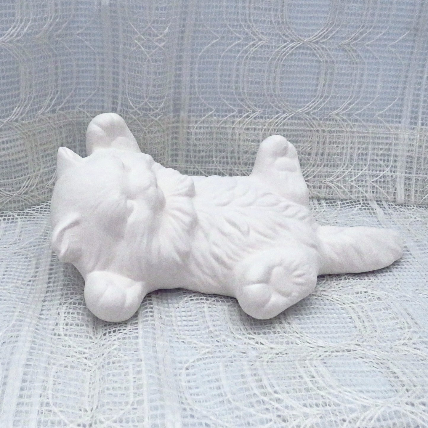 Paintable ceramic kitten on its back lying on a table with a lacy ecru table cloth.