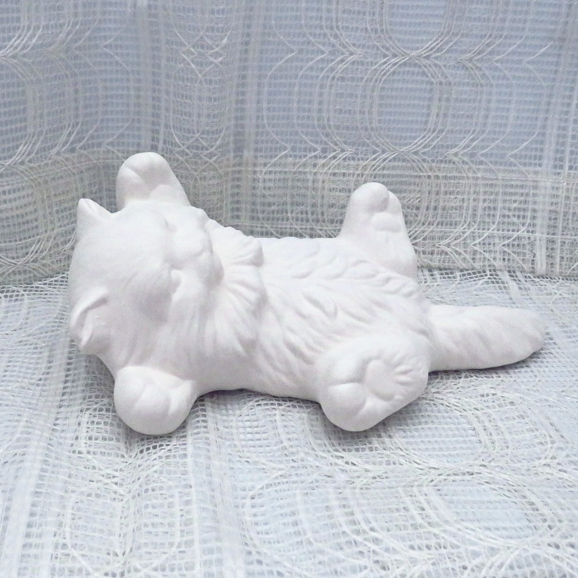 Paintable ceramic kitten on its back lying on a table with a lacy ecru  table cloth.
