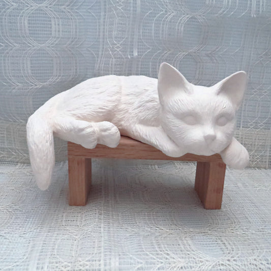 handmade ready to paint ceramic cat lying on a wooden pedestal with an ecru curtain and table cloth.  The cat's tail hangs below the surface of the pedestal, as does his frong paws and chin
