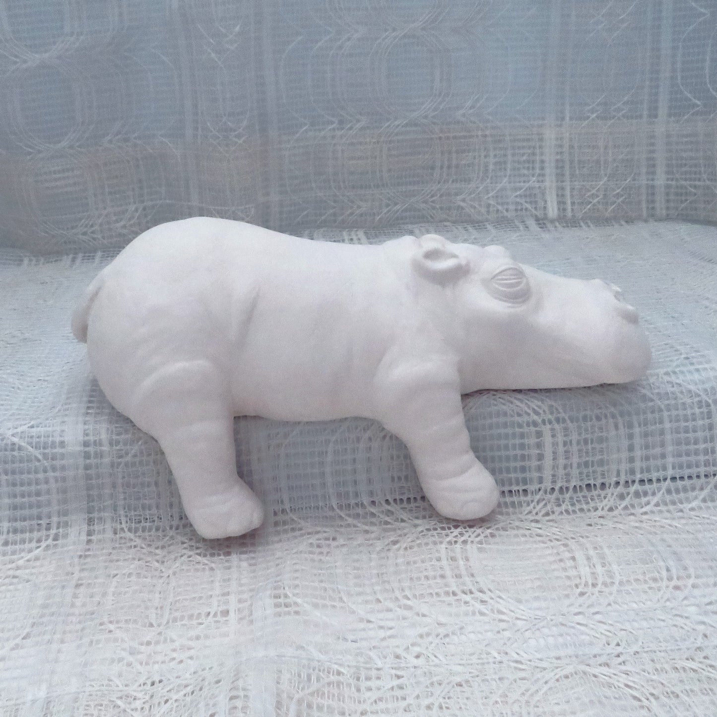 Large Ready to paint ceramic hippo figurine sitting on a shelf with its right legs hanging off. There is an ecru lacy curtain behind and under it.