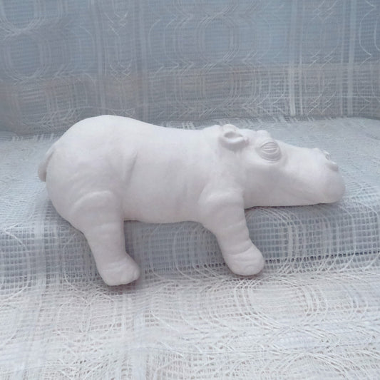 Large Ready to paint ceramic hippo figurine sitting on a shelf with its right legs hanging off.  There is an ecru lacy curtain behind and under it.