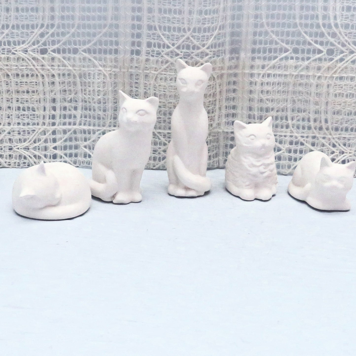 Set of 5 ready to paint small ceramic cats on a blue table with an off white lacy fabric behind them. They are positioned so the tallest ones are in the center and the shortest ones at the ends