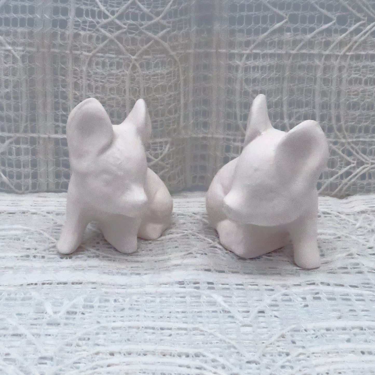 Set of 2 handmade ready to paint small pigs sitting looking toward each other. They are sitting on a lacy background.