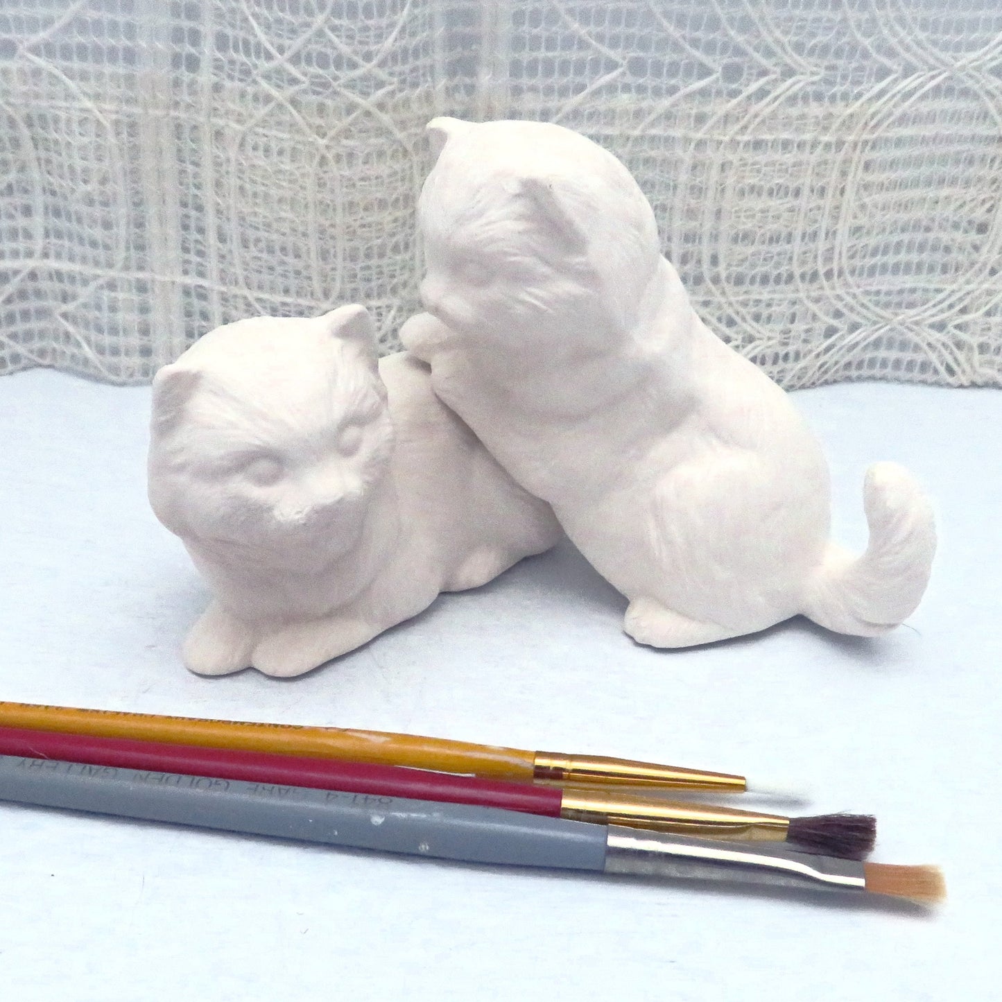2 paintable ceramic cats, one lying down, one jumping on the other one. They are on a blue table and an ecru curtain behind it. There are 3 paint brushes on the table in front of them.