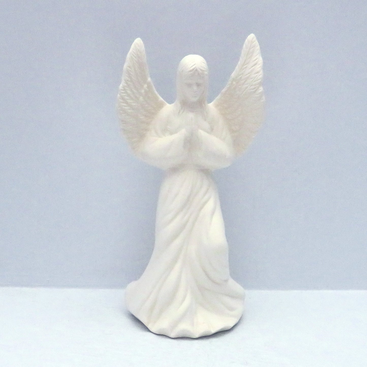 Unpainted ceramic praying angel figurine on a pale blue surface. Her wings are outstretched and her head is slightly bowed.
