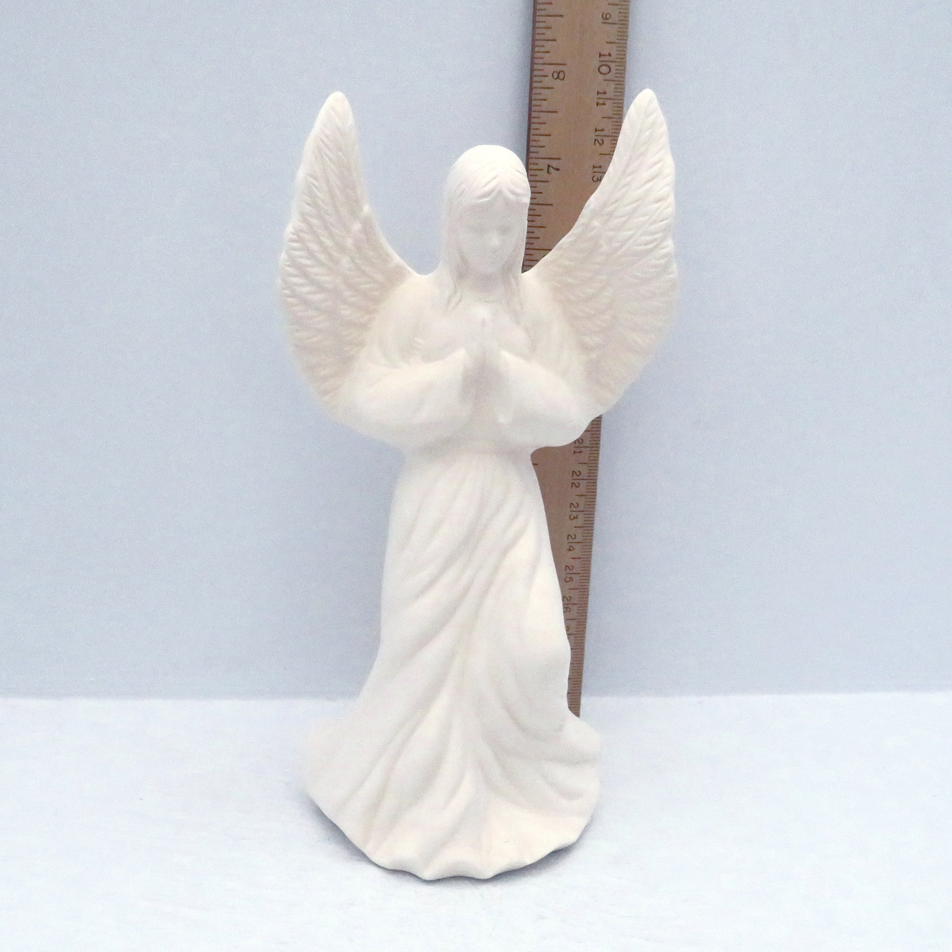 Unpainted ceramic bisque figurine standing in front of a ruler showing her height to be approximately 8 inches tall at the top of the wing