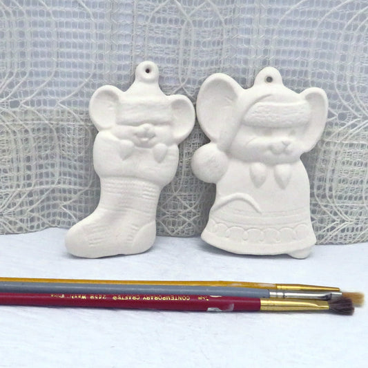 2 unpainted ceramic Christmas tree ornaments featuring a little mouse in a stocking and climbing over a bell on a pale blue table with 3 paint brushes in front.