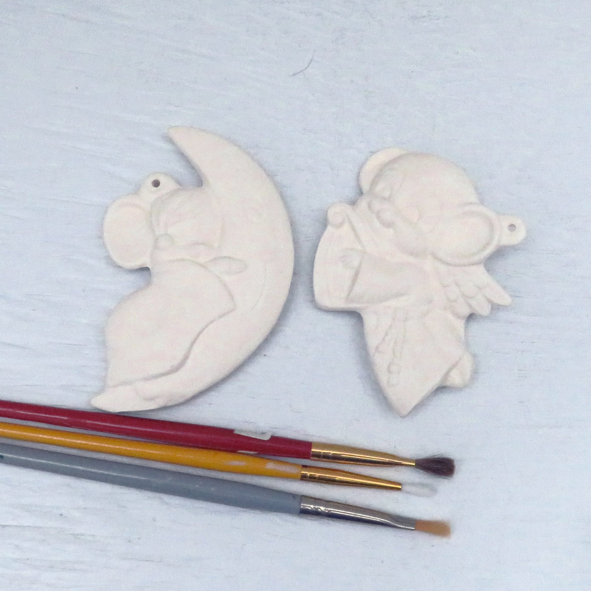 Handmade paintable ceramic Christmas tree ornaments showing a mouse on a crescent moon and an angel mouse with a harp.  There are 3 paint brushes , not included in the set on a pale blue background.