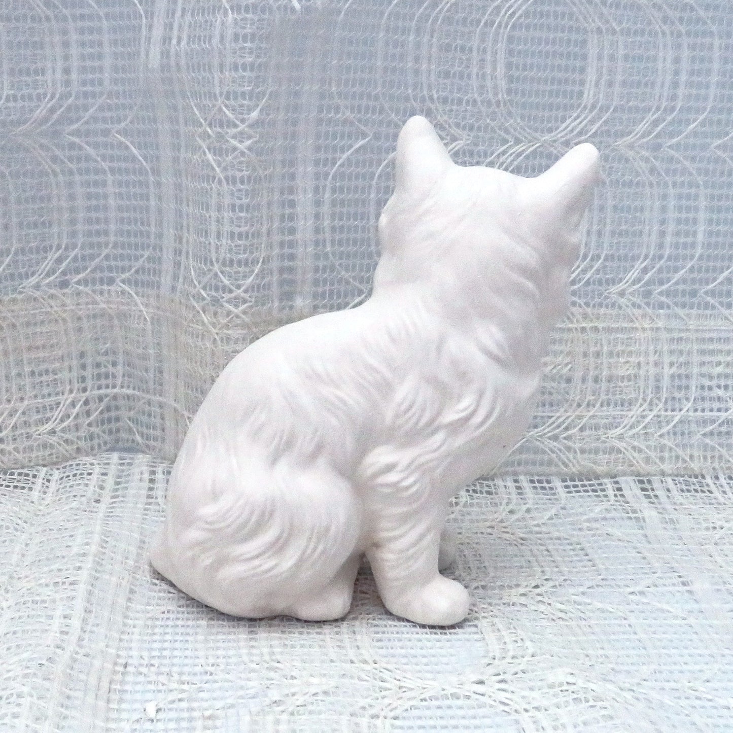 Paintable ceramic persian cat sitting on a table with a lacy ecru table cloth and curtain. This photo shows the cat facing away from the camera.