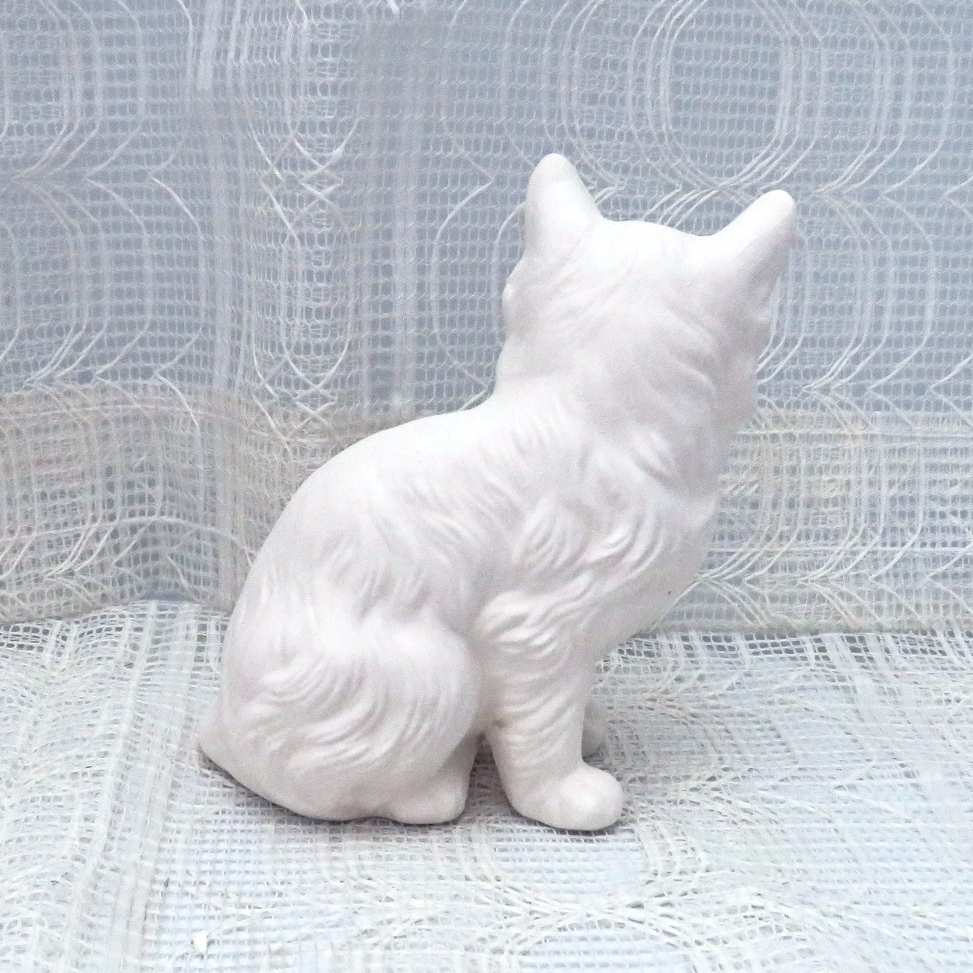 Paintable ceramic persian cat sitting on a table with a lacy ecru table cloth and curtain.  This photo shows the cat facing away from the camera.