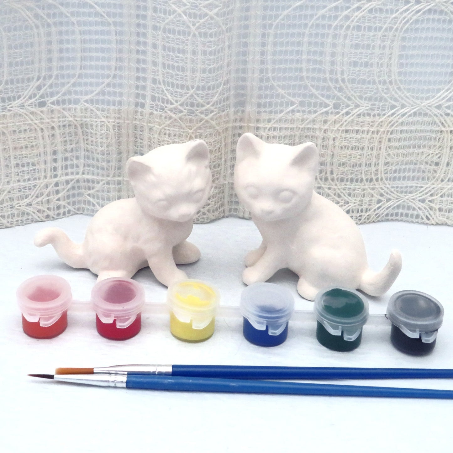 2 unpainted ceramic cat statues sitting on a pale blue table with an ecru lacy curtain behind them. The cats are sitting with their fronts facing each other, and their heads turned to face the camera. There is a set of acrylic paints in plastic tubs and snap lids and 2 paint brushes in front of them.