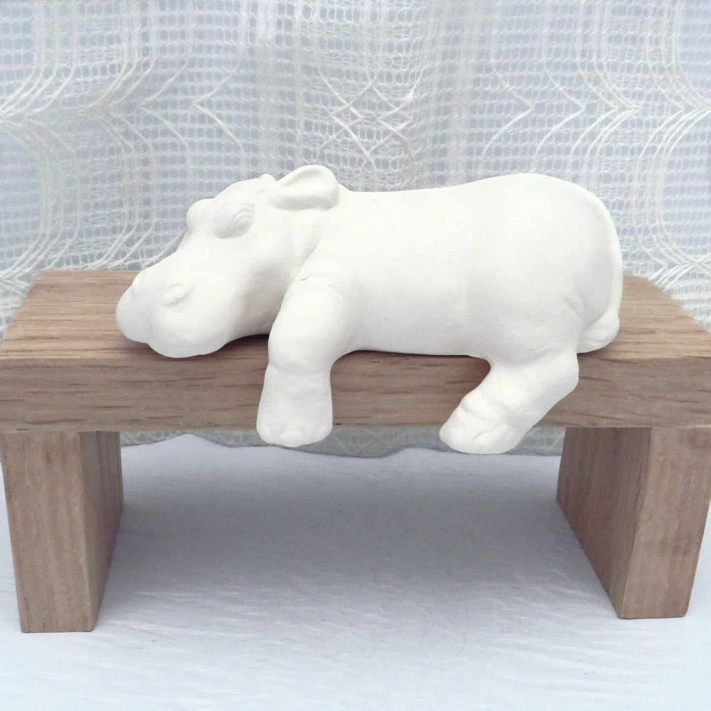 handmade ready to paint ceramic hippo figurine lying on a brown wooden pedestal. This hippo is a shelf sitter. Its chin, left front and rear leg are hanging over the shelf.