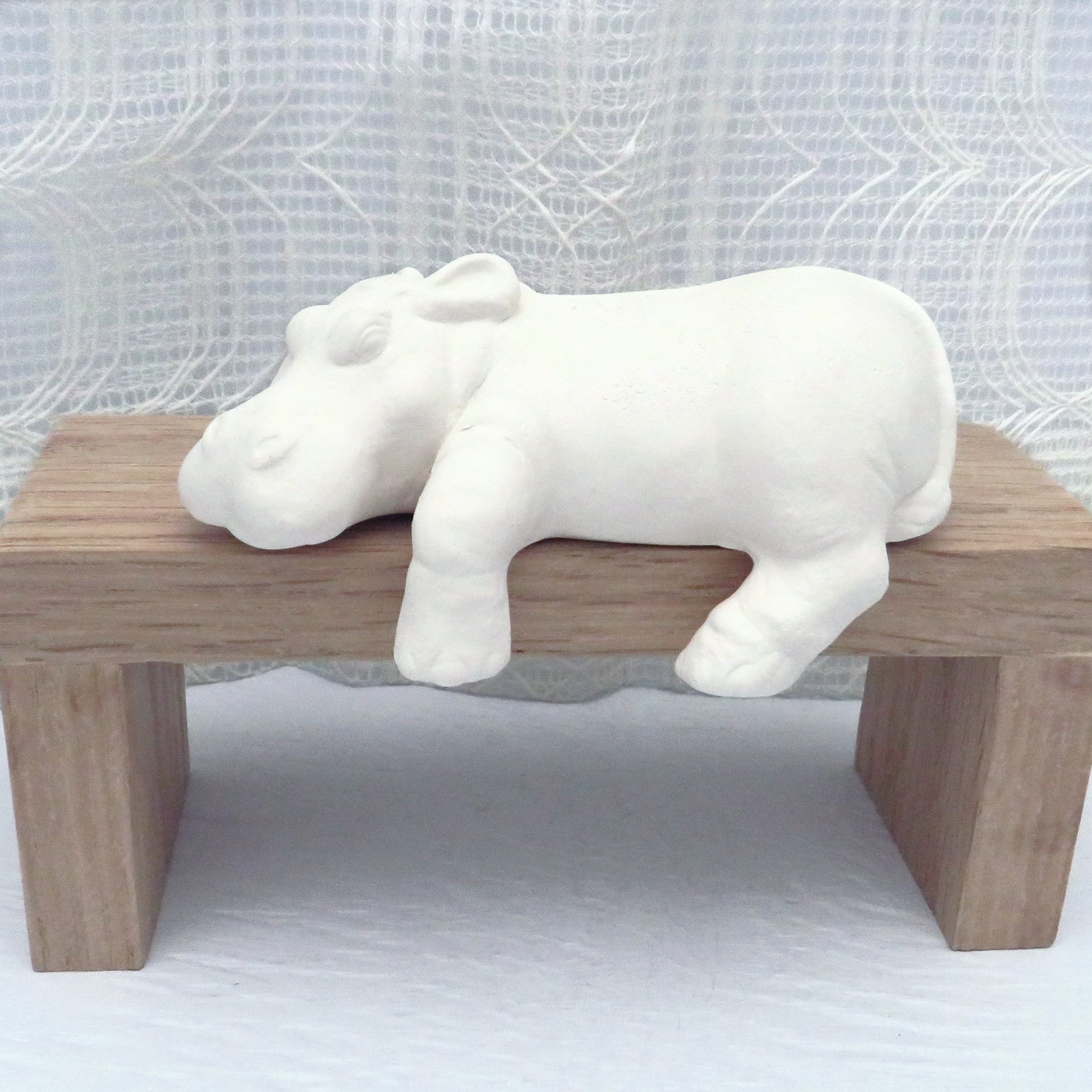 handmade ready to paint ceramic hippo figurine lying on a brown wooden pedestal.  This hippo is a shelf sitter.  Its chin, left front and rear leg are hanging over the shelf.