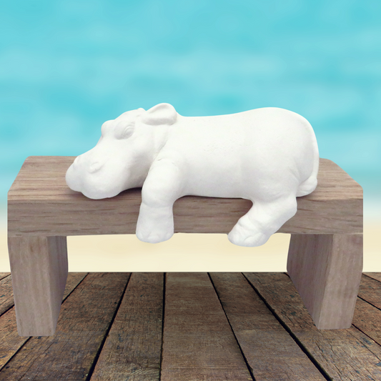 Small unpainted ceramic shelf hippo on a wooden pedestal on a rustic wood table by the ocean.   The hippo's left legs are hanging over the pedestal