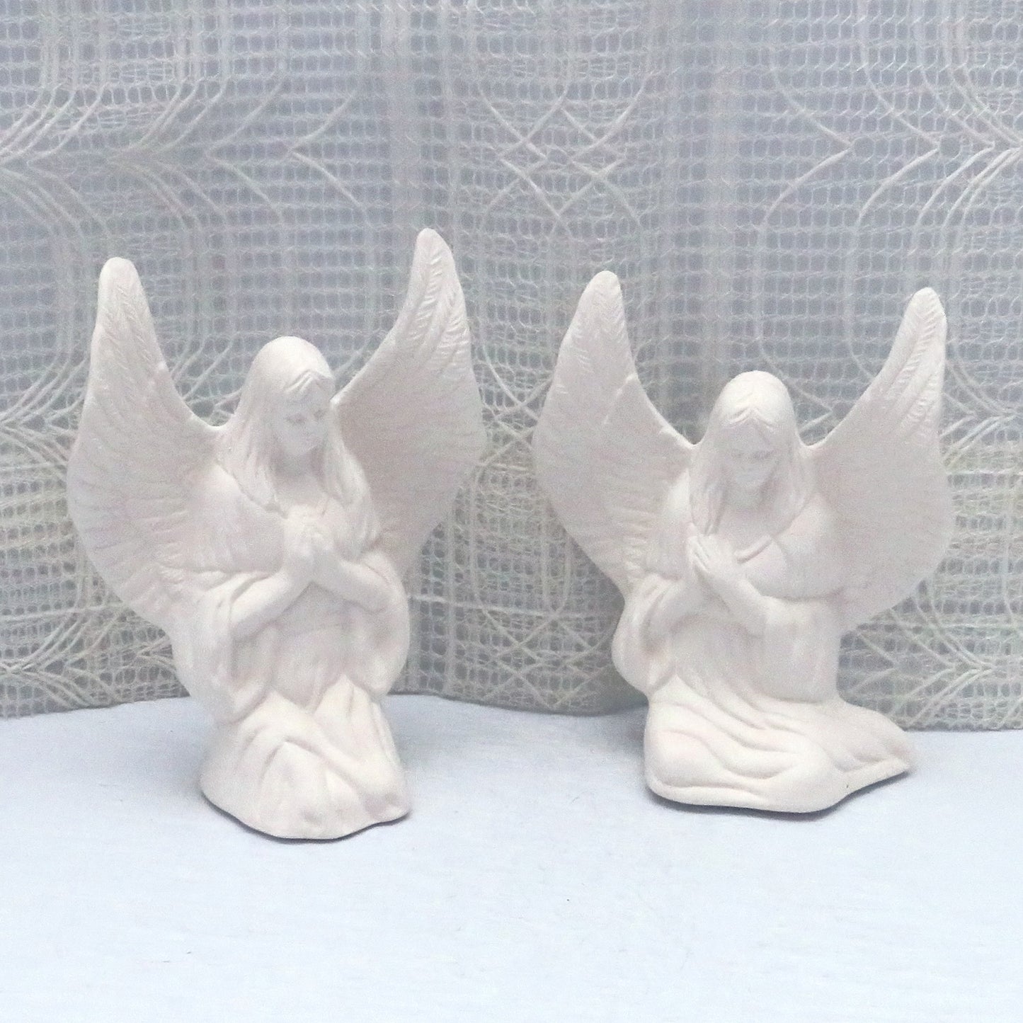 Handmade ready to paint ceramic angek statues on a blue tablle with an off white lacy curtain behind. The angels are facing forward