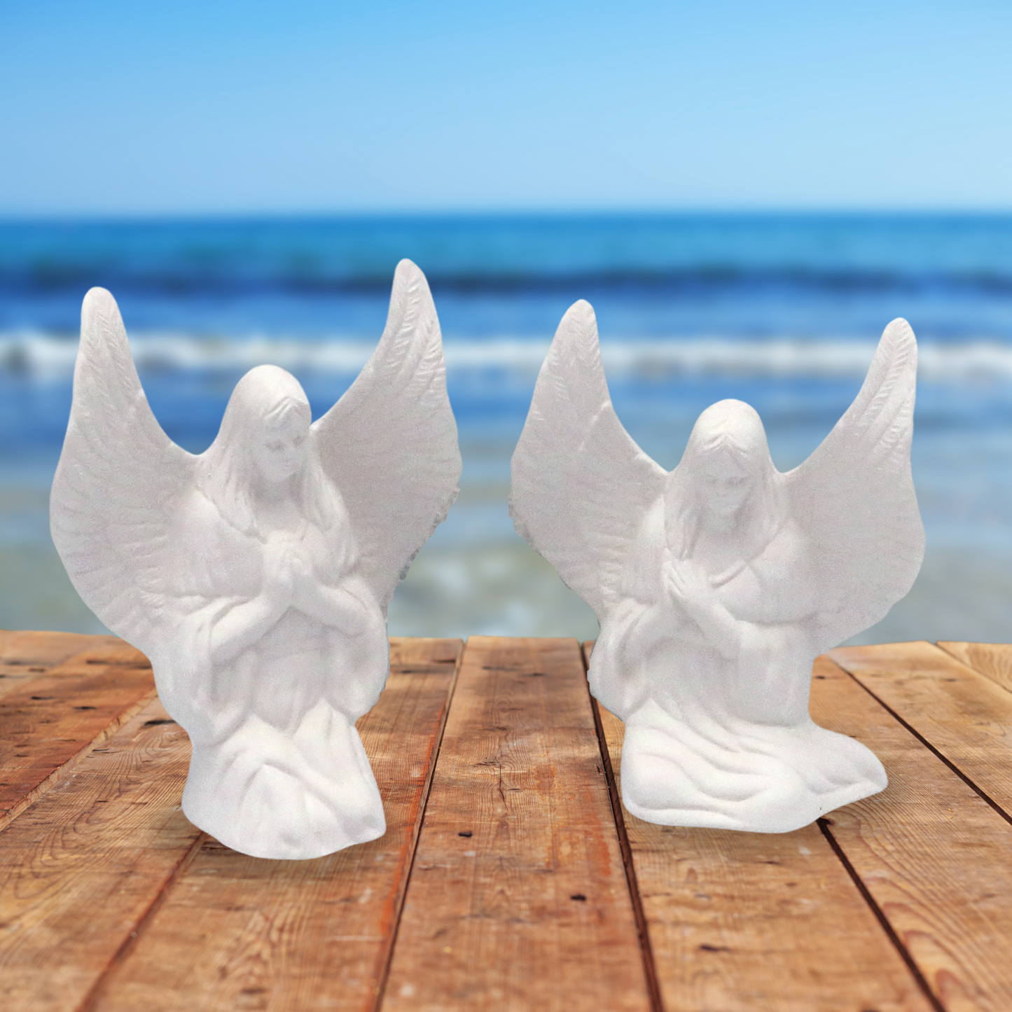 Set of 2 unpainted ceramic angel figurines sitting on a wood table by the ocean. Both of the angels have their wings oustretched. One is sitting side sitting, one is kneeling.