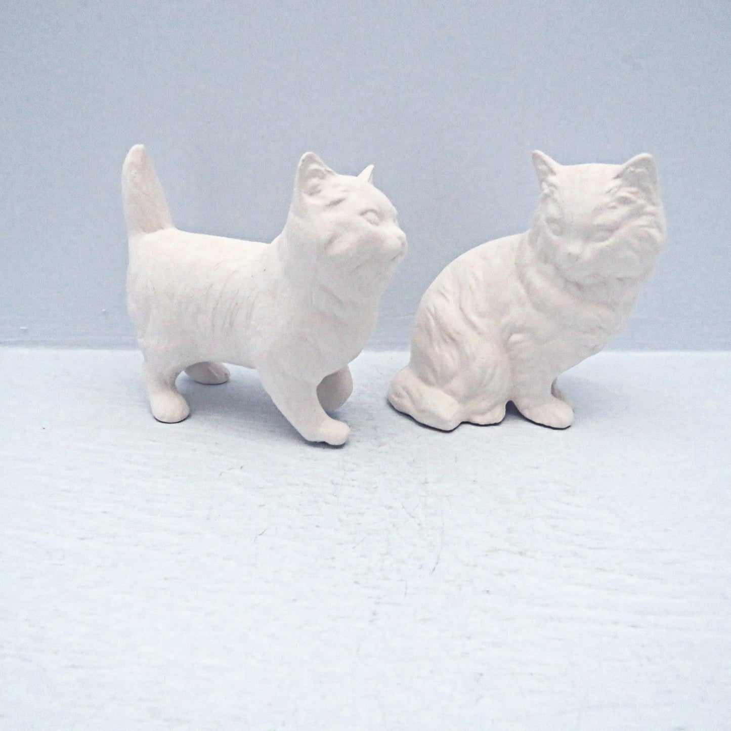 2 ready to paint ceramic cat figurines. The one on the left is standng and facing the right. The one on the left is sitting with its body toward the right, and its head facing the camera The standing cat has its tail pointing up. They are on a pale blue surface.