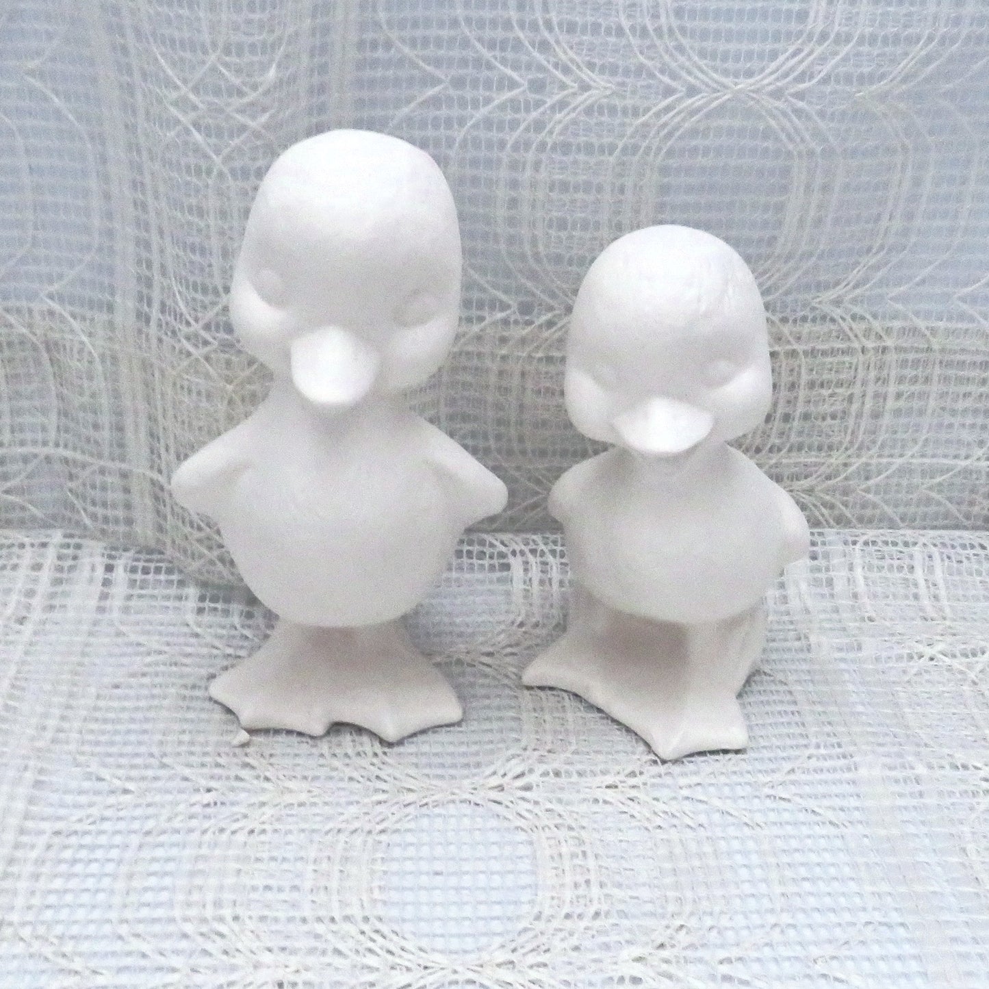 Set of 2 ready to paint ceramic duck figurines standing on a table with a lacy ecru table clote. The ducks are facing forward. Ond is shorter than the other and the shorter one has its mouth open..