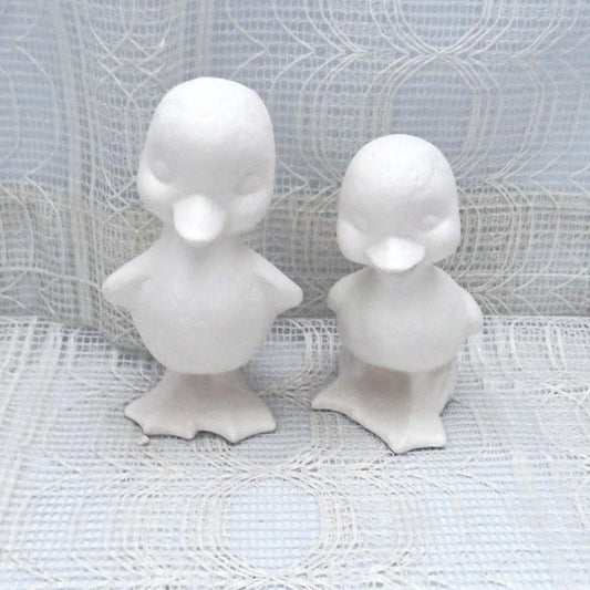Set of 2 ready to paint ceramic duck figurines standing on a table with a lacy ecru table clote.  The ducks are facing forward.  Ond is shorter than the other and the shorter one has its mouth open..
