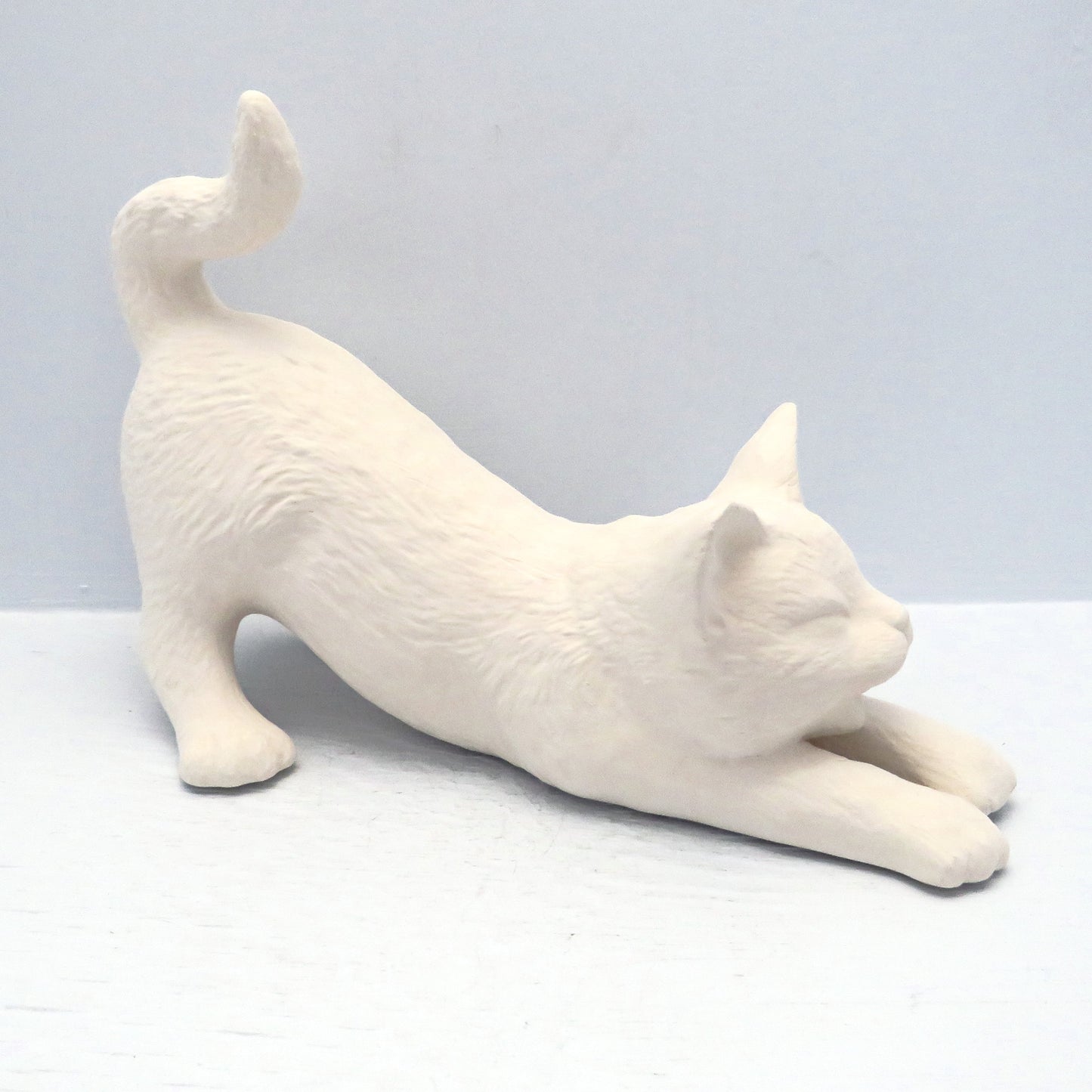 Angle view of ready to paint ceramic stretching cat on a pale blue background. The tale and rear of the cat are up, the head is down and the front legs are stretched forward.