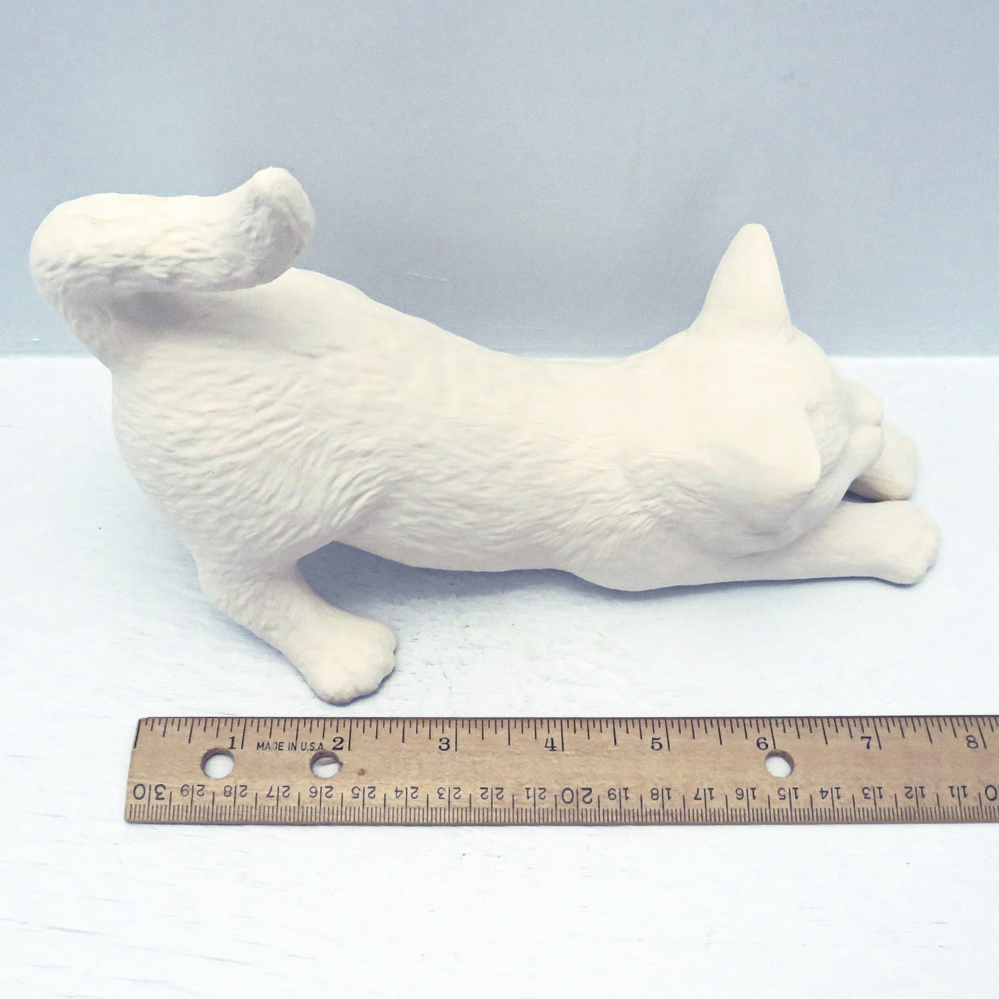 Handmade ready to paint ceramic stretching cat with head to the right. It s on a pale blue surface.