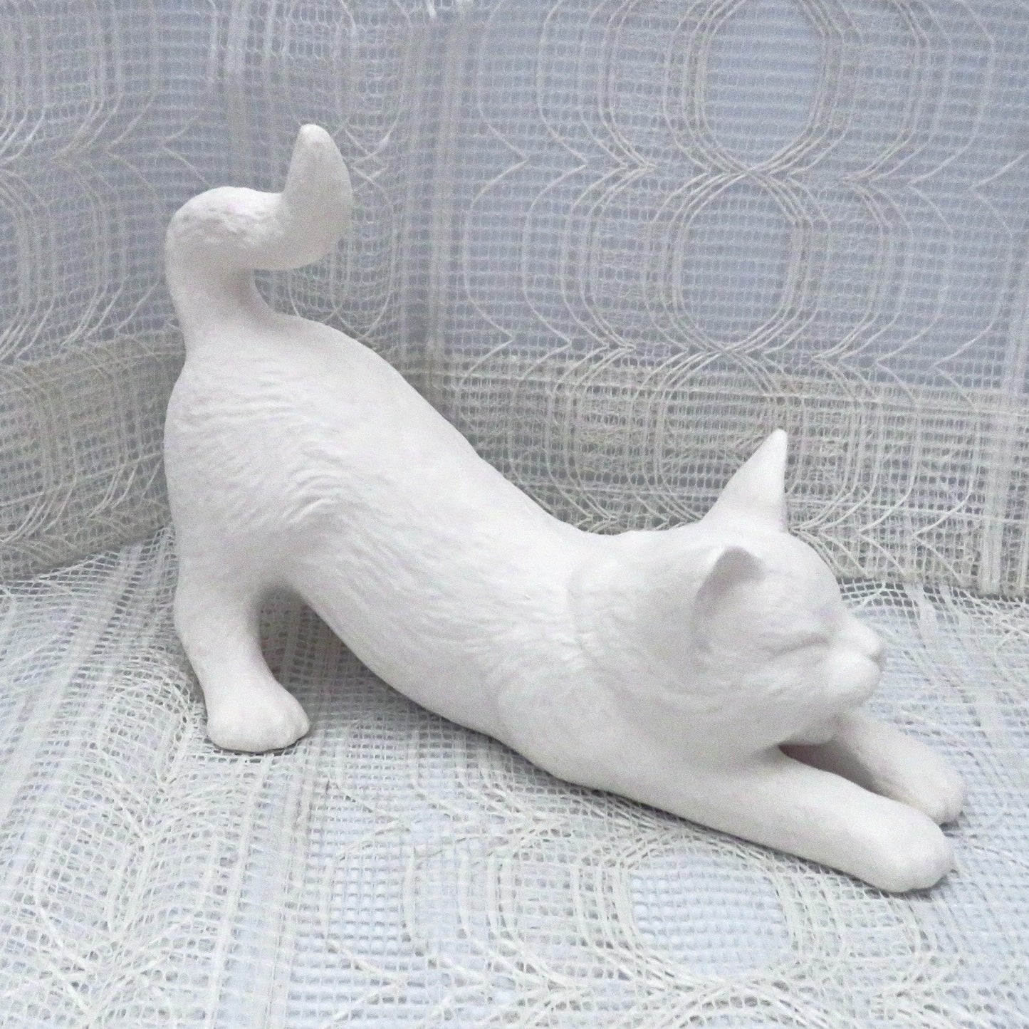 Paintable ceramic cat stretching on a table with a lacy ecru table cloth with an ecru curtain behind it.