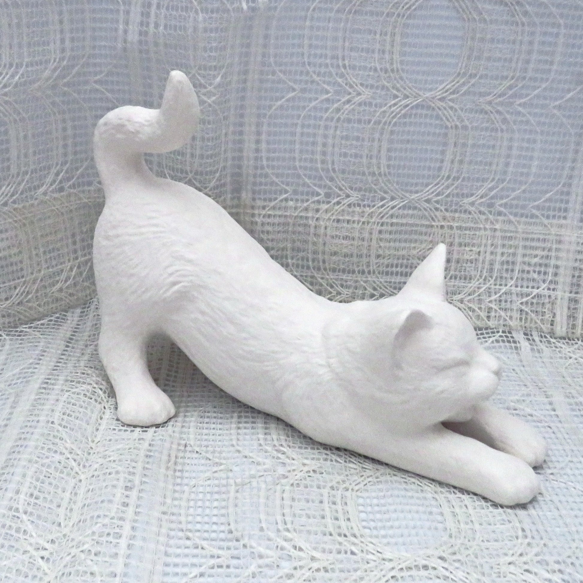 Paintable ceramic cat stretching on a table with a lacy ecru table cloth with an ecru curtain behind it.